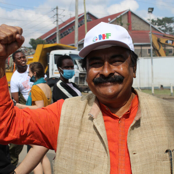 A man of colour is standing outside smiling with his right fist in the air. He is wearing a white hat that reads "NHF."